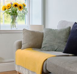 A sofa with cushions and a yellow throw next to a window with sunflowers in a vase at Cove Cottage in Porthgwarra