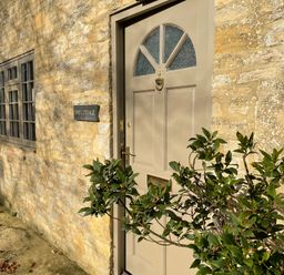 An exterior view of a stone cottage with a beige wooden door a window and a small plant nearby at Omas Cottage Moreton-In-Marsh
