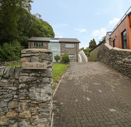 A driveway leading to a stone house with a lawn and a wooden sign saying Tan Twr next to a modern building at Tan Twr in Llanfairpwll