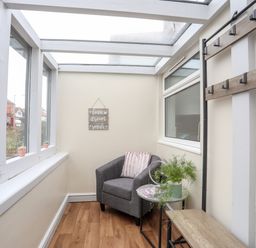 A small sunroom with a grey armchair a small table with a plant a coat rack and windows at Beeches Bach in Prestatyn