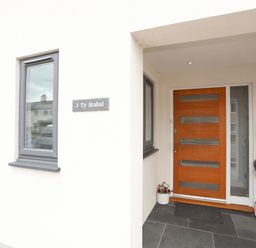 An entrance area with a wooden front door a gray window two potted plants a mailbox and a nameplate saying 3 Ty Stabal in Pwllheli