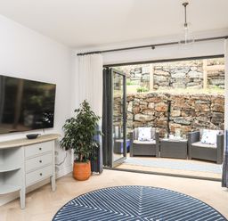 A living area with a wall-mounted television a sideboard plant and bi-fold doors leading to an outdoor seating area with chairs a table and a stone wall at 3 Ty Stabal in Pwllheli