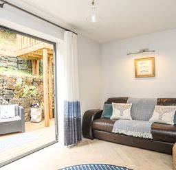 A living room with a dark leather sofa and cushions next to a sliding glass door opening to a patio with outdoor chairs at 3 Ty Stabal in Pwllheli
