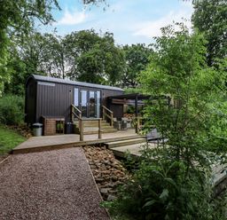 A wooden hut with steps and outdoor seating surrounded by trees and plants at The Shepherds Hut at Bridge Lake Farm & Fishery in Chacombe