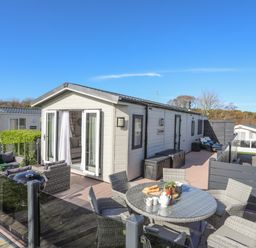 An outdoor patio area with a table and chairs a barbecue and seating in front of a beige modular home at 97 Crugan in Llanbedrog