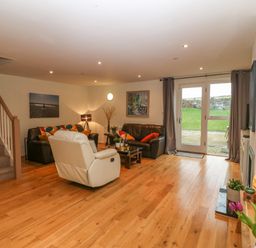 A living room with brown sofas a white armchair wooden floor and glass doors leading outside at Barn End 9 Stad Clynnog in Newborough