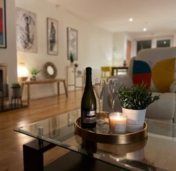 A living room with a bottle, glasses, candle, and plant on a glass table near an armchair with colorful pillows and a fireplace at Barn End, 9 Stad Clynnog in Newborough