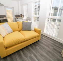 A yellow sofa with pillows in a room with large windows and a kitchen and dining area visible at Perran Pearl A in Perranporth