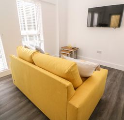 A yellow sofa with cushions in a living room with a wall-mounted TV and a small wooden nesting table at Perran Pearl A in Perranporth