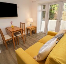 A room with a yellow sofa with pillows a small wooden dining table with two chairs a nested side table with a lamp and glass doors leading outside at Perran Pearl B in Perranporth