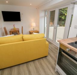 A living area with a yellow sofa, dining table, wall-mounted TV, lamp, and kitchen oven with glass doors at Perran Pearl B in Perranporth
