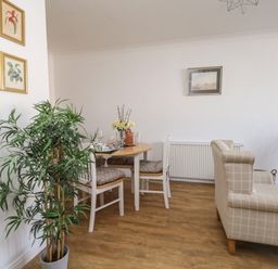 A dining area with a round table set with glassware and white chairs beside a beige checkered sofa in a room with wooden flooring and wall art at Beach Side in West Bay