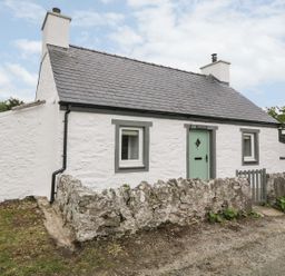 A white stone cottage with two chimneys and green door surrounded by a low stone wall at Rhytty Bach in Ty Croes near Rhosneigr