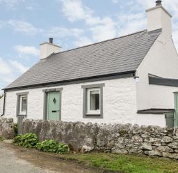 A white stone cottage with green doors and windows behind a low stone wall at Rhytty Bach in Ty Croes near Rhosneigr