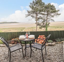 An outdoor patio area with a small round table and two chairs overlooking a sandy beach and grassy area at Oversands Cottage in Grange-Over-Sands