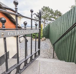 A metal gate with Oversands Cottage sign next to stone steps and gravel path at Oversands Cottage in Grange-Over-Sands