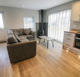 A living area with a grey corner sofa, coffee table, wood stove, and kitchen with wooden floor at White Stones in Goadsbarrow near Roose