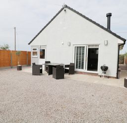 A backyard with a gravel ground a white house with a sliding door outdoor dining table with chairs and a barbecue at White Stones in Goadsbarrow near Roose
