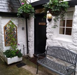 A black door with the number 5 and a bench in front of a white stone wall with a window and hanging plants at Fox Hat Cottage in Chagford