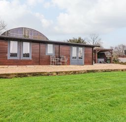 A wooden cabin with gray window frames and doors on a gravel patio with chairs and table on a lawn at Westfield Lodge in Terrington near Sheriff Hutton