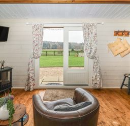 A living room with a leather chair a round wooden table with a plant floral curtains over glass doors opening to a green field at Westfield Lodge in Terrington near Sheriff Hutton