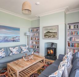 A living room with two blue sofas, a wooden coffee table, bookshelves, a fireplace with a painting above it, and a floor lamp at Bryn Henllan in Criccieth