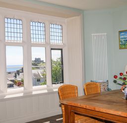 A dining area with a wooden table and wicker chairs next to large leaded windows showing a view of rooftops and a castle at Bryn Henllan in Criccieth