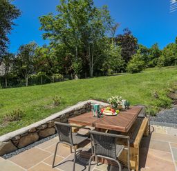 An outdoor patio area with a wooden table and chairs next to a grassy slope with trees at Bryn Henllan in Criccieth