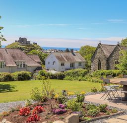 A garden with flowers and a patio table with chairs overlooking houses a stone building and a castle on a hill in Criccieth