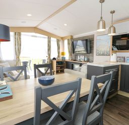 A dining table with chairs next to a kitchen and a living area with a sofa and television at Trewan Lodge in Bryngwran near Rhosneigr
