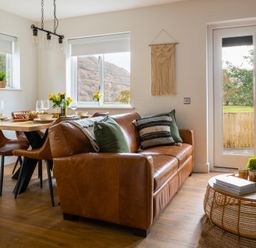 A dining area with a wooden table, four chairs, a brown leather sofa, round coffee table with cups and books at Sygun in Beddgelert
