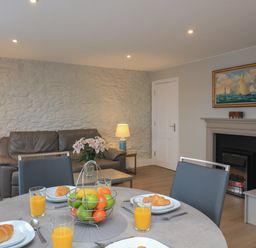 A dining table with plates of croissants and glasses of orange juice in a living room with a brown sofa, lamps, a fireplace, and a painting at Queens View in Dartmouth
