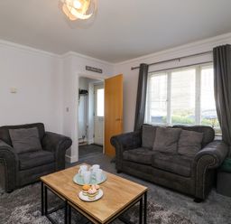 A living room with a sofa and armchair a coffee table with mugs and cupcakes a large window with curtains at Anchors Away in Weymouth