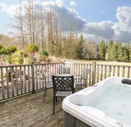 A wooden deck with a hot tub and metal chairs around a table overlooking trees and plants at The Lodge in Dol-y-Gaer near Pontsticill
