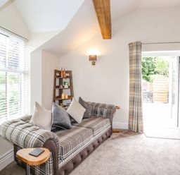 A living room with a plaid sofa and pillows a wooden shelf with decor and candles and a door with plaid curtains at Chestnuts Cottage in Windermere