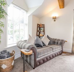 A sitting room with a plaid sofa cushions a basket with blankets a wooden shelf with candles and a window with blinds and curtains at Chestnuts Cottage in Windermere