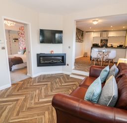 A living room with a brown leather sofa and pillows a wall mounted TV a fireplace an open doorway to a bedroom and an open kitchen and dining area at The Chalet in Benllech