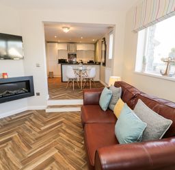 A living room with a brown leather sofa and cushions a wall-mounted tv and a fireplace with a view to a dining area and kitchen at The Chalet in Benllech