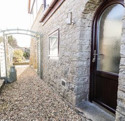 A stone wall with an arched wooden door and frosted window beside a narrow gravel path and a white wooden fence with an archway over the path at The Chalet in Benllech