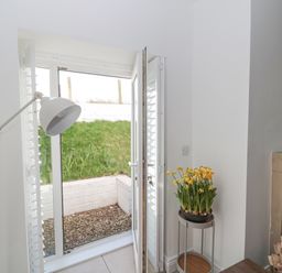 A room with an open glass door showing a small outdoor area with grass and white brick wall and a plant with yellow flowers on a stand next to a fireplace at Little Haven in Bamburgh