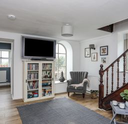 A living room with a mounted TV above a cabinet with DVDs and board games a grey armchair under framed pictures and a wooden staircase with a window and plant at Bethania in Llangoed near Beaumaris