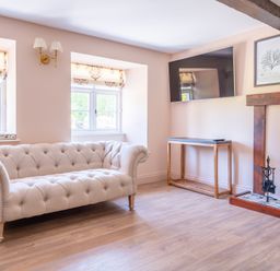 A living room with a beige tufted sofa two windows a wall-mounted TV a wooden console table and a fireplace at Cunsey Mill House in Cunsey near Hawkshead
