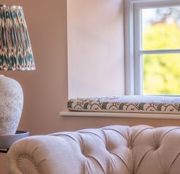 A room corner with a patterned lampshade on a stone base lamp a cushioned beige sofa and a window with a floral cushion on the sill at Cunsey Mill House Cunsey near Hawkshead
