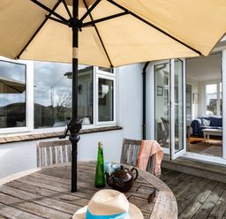 An outdoor patio with a round wooden table and chairs under a beige umbrella at Croft Elms in Trebetherick near Polzeath