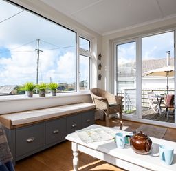 A sunroom with wicker chairs a bench with cushions a white coffee table and large windows overlooking an outdoor patio at Croft Elms in Trebetherick near Polzeath