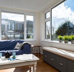 A sitting area with a blue covered sofa two cushions a white coffee table with a teapot two mugs and maps a bench with storage and potted plants on the windowsill at Croft Elms in Trebetherick near Polzeath