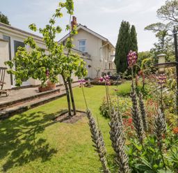 A garden with grass, flowering plants, small tree, wooden chairs, and a house in the background at Bryn Offa cottage in Holywell