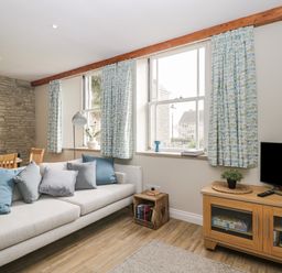 A living room with a sofa and cushions next to three windows with patterned curtains and a wooden TV stand with a plant at The Old Haberdashery in Swanage