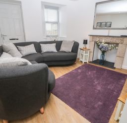 A living room with a curved gray sofa a purple rug and flowers in a fireplace at Rose Bank Cottage in St Just