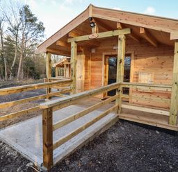 A wooden cabin with a small porch and railing in a wooded area at Buttercup Lodge in Theberton near Leiston